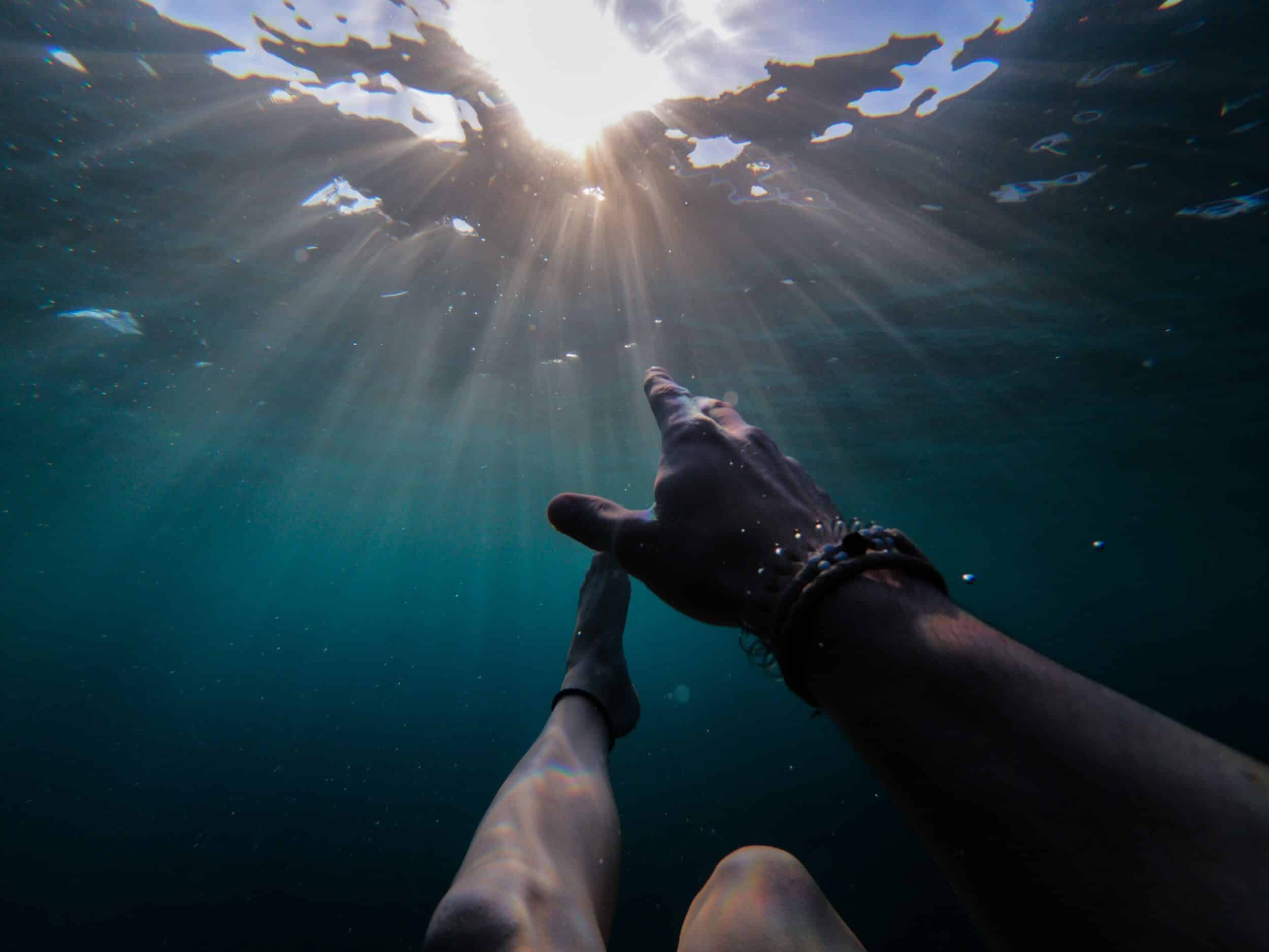 An underwater view of a hand reaching up toward sunlight streaming through the water