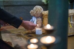 A hand placing a tissue beside a teddy bear and candle at a memorial